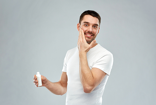 beauty, skin care, body care and people concept - smiling young man applying cream or lotion to face over gray background