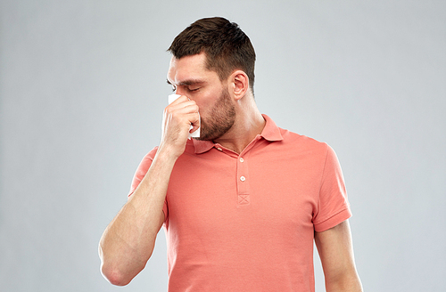 people, healthcare, rhinitis, cold and allergy concept - sick man with paper napkin blowing nose over gray background