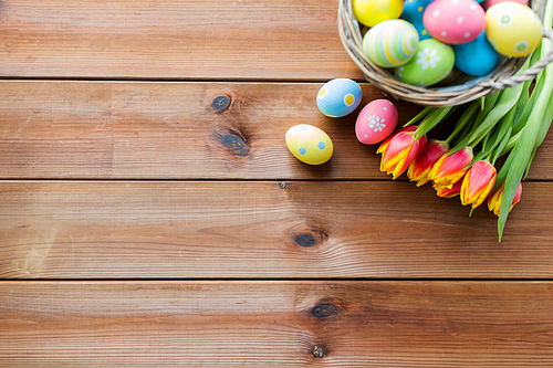 easter, holidays, tradition and object concept - close up of colored easter eggs in basket and tulip flowers on wooden table with copy space