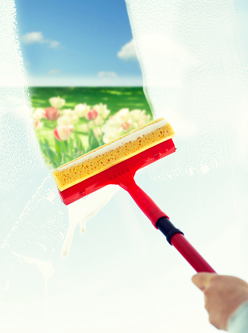 people, housework and housekeeping concept -close up of hand cleaning window glass with sponge mop and foam over blue sky and flower field background