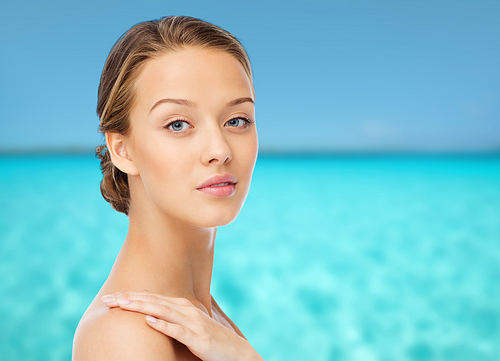beauty, people, body care and health concept - smiling young woman face and hand on bare shoulder over blue sea and sky background