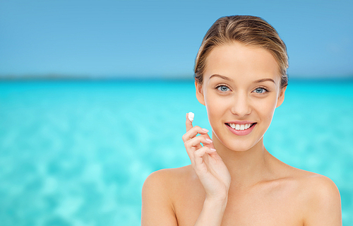 beauty, people, cosmetics, skincare and health concept - happy smiling young woman applying cream to her face over blue sea and sky background