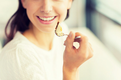 food, dessert, people and lifestyle concept - close up of smiling young woman holding fork and eating cake at cafe or home