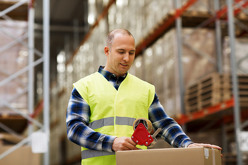 logistic, delivery, shipment, people and export concept - happy man in safety vest packing box or parcel with scotch tape at warehouse or  storage