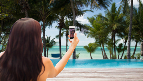 summer, travel, technology and people concept - close up of  young woman taking selfie with smartphone over tropical beach with palms and swimming pool background