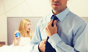 people, business, morning and work concept - close up of man in shirt dressing up and adjusting tie on neck over bedroom with woman holding alarm clock background