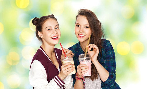 people, friends, teens and friendship concept - happy smiling pretty teenage girls drinking milk shakes and with straw over green holidays lights background