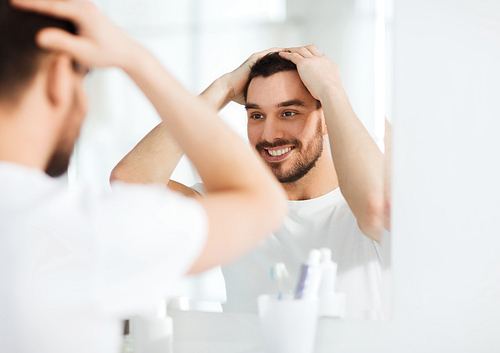 beauty, hygiene and people concept - smiling young man looking to mirror and styling hair at home bathroom