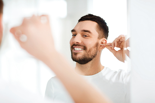 beauty, hygiene and people concept - smiling young man cleaning ear with cotton swab and looking to mirror at home bathroom