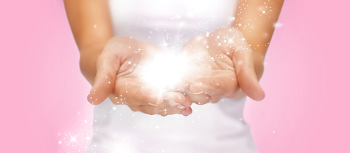 people and magic concept - close up of twinkles or fairy dust on female cupped hands over pink background