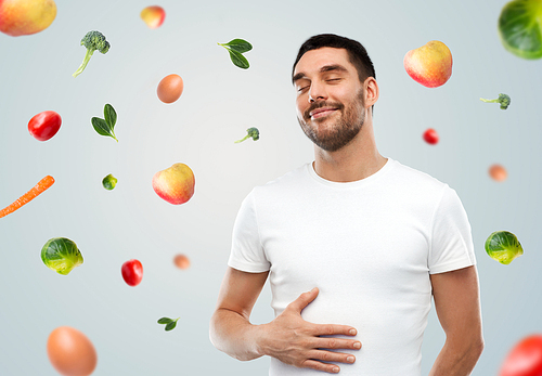 food, healthy eating, diet and people concept - happy full man touching his tummy over gray background with falling fruits