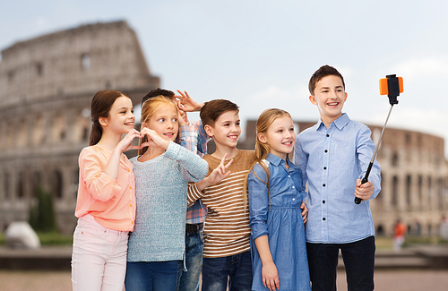childhood, travel, tourism, technology and people concept - happy children talking picture by smartphone on selfie stick over coliseum in rome