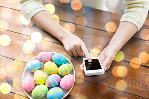 easter, holidays, tradition, technology and people concept - close up of woman hands with colored easter eggs on plate and smartphone over holidays lights