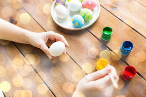 easter, holidays, tradition and people concept - close up of woman hands coloring easter eggs with colors and brush over holidays lights