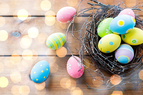 easter, holidays, tradition and object concept - close up of colored easter eggs in nest on wooden surface over holidays lights