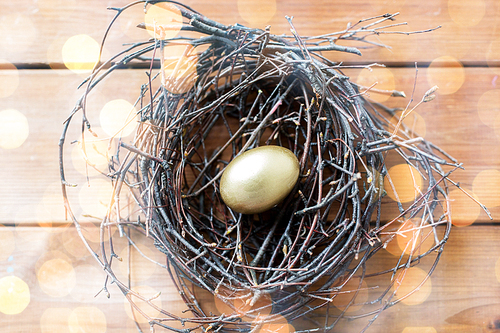 easter, holidays, tradition and object concept - close up of golden easter egg in nest on wooden surface over holidays lights