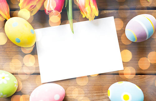 easter, holidays, tradition and object concept - close up of colored easter eggs, tulip flowers and blank white paper card on wooden surface with copy space over holidays lights