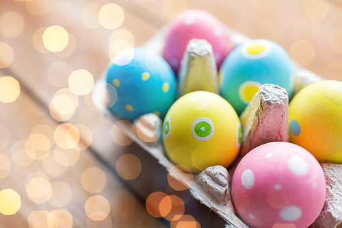 easter, holidays, tradition and object concept - close up of colored easter eggs in egg box or carton wooden surface over holidays lights