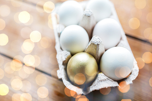 easter, food, cooking and object concept - close up of white and golden eggs in egg box or carton wooden surface over holidays lights