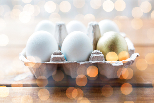 easter, food, cooking and object concept - close up of white and golden eggs in egg box or carton wooden surface over holidays lights