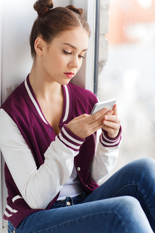 people, technology and teens concept - sad unhappy pretty teenage girl sitting on windowsill with smartphone and texting