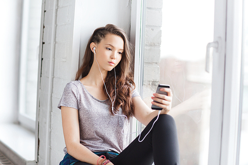 people, technology and teens concept - sad pretty teenage girl sitting on windowsill with smartphone and earphones listening to music