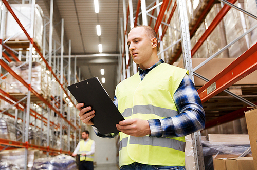 wholesale, logistic, people and export concept - man with clipboard in reflective safety vest at warehouse