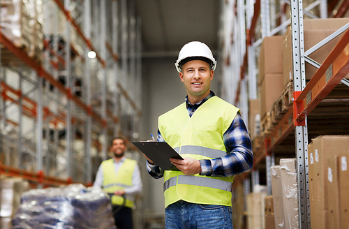 wholesale, logistic, people and export concept - man with clipboard in reflective safety vest at warehouse