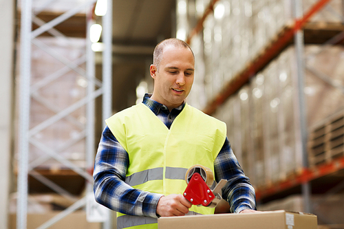 logistic, delivery, shipment, people and export concept - happy man in safety vest packing box or parcel with scotch tape at warehouse or mail storage