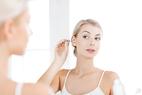 beauty, hygiene and people concept - young woman cleaning ear with cotton swab and looking to mirror at home bathroom