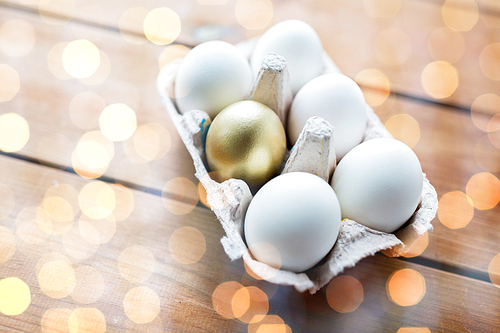 easter, food, cooking and object concept - close up of white and golden eggs in egg box or carton wooden surface over lights