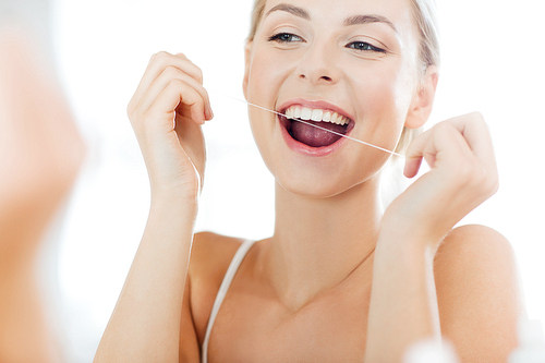 health care, dental hygiene, people and beauty concept - smiling young woman with floss cleaning teeth and looking to mirror at home bathroom