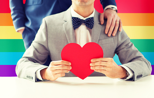people, homosexuality, same-sex marriage, valentines day and love concept - close up of happy married male gay couple with red paper heart shape on wedding over rainbow flag background