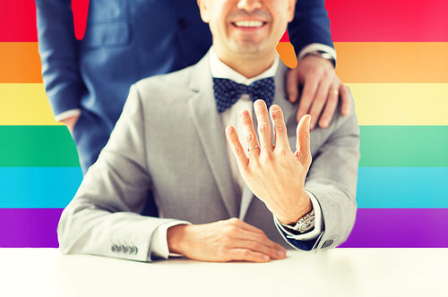 people, celebration, homosexuality, same-sex marriage and love concept - close up of male gay couple with wedding rings on putting hand on shoulder over rainbow flag background