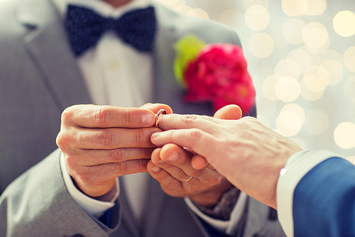 people, homosexuality, same-sex marriage and love concept - close up of happy male gay couple hands putting wedding ring on lights background