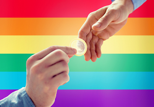 people, homosexuality, safe sex, sexual education and charity concept - close up of happy male gay couple hands giving condom over rainbow flag background