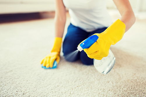 people, housework and housekeeping concept - close up of woman in rubber gloves with cloth and derergent spray cleaning carpet at home