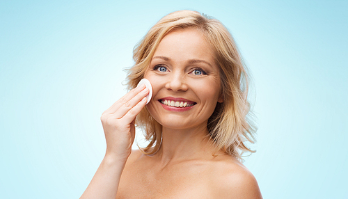 beauty, people and skincare concept - happy middle aged woman cleaning face and removing make up with cotton pad over blue background