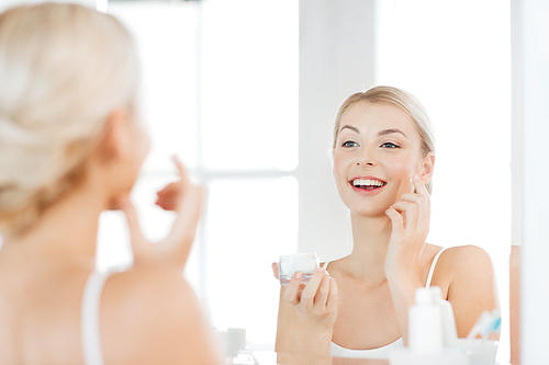 beauty, skin care and people concept - smiling young woman applying cream to face and looking to mirror at home bathroom