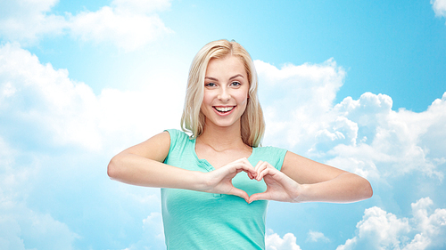 gesture and people concept - smiling young woman or teenage girl showing heart shape made of fingers over blue sky and clouds background