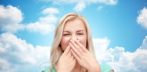emotions, expressions, embarrassment, and people concept - young woman or teenage girl wrinkling and closing her nose of unpleasant smell over blue sky and clouds background
