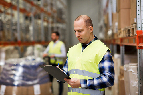 wholesale, logistic, people and export concept - man with clipboard in reflective safety vest at warehouse