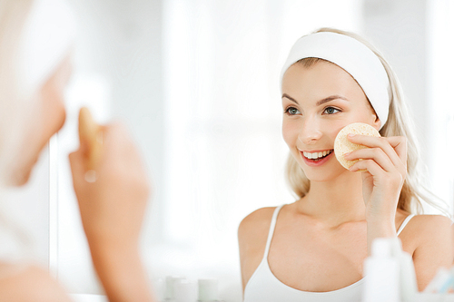 beauty, skin care and people concept - smiling young woman washing her face with facial cleansing sponge at bathroom