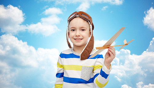 childhood, fashion and people concept - happy smiling little boy in aviator hat playing with wooden airplane over blue sky and clouds background