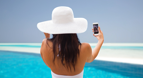 lifestyle, leisure, summer, technology and people concept - smiling young woman or teenage girl in sun hat taking selfie with smartphone over beach and swimming pool background