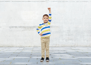 childhood, power, gesture and people concept - happy smiling little boy with raised hand over urban concrete background