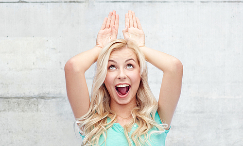 fun, expressions, and people concept - happy smiling young woman making bunny ears over gray concrete wall background
