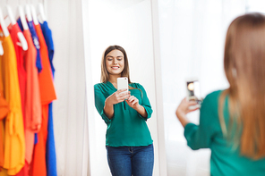 clothing, fashion, style, technology and people concept - happy woman with smartphone taking mirror selfie at home wardrobe