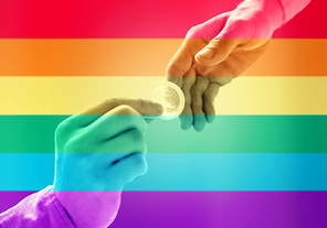 people, homosexuality, safe sex, sexual education and charity concept - close up of male gay couple hands giving condom over rainbow flag stripes background