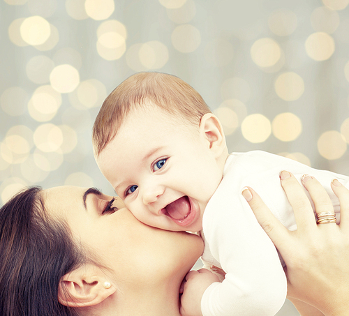 people, family, motherhood and children concept - happy mother hugging adorable baby over holidays lights background